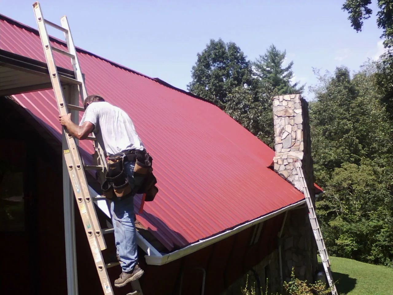 Roofer on a ladder beside a red metal roof and stone chimney — Soffit & Fascia Repair by Roofing Brothers in Lake Lure